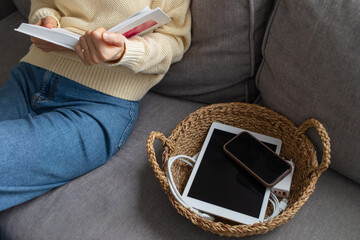 Digital detox concept from woman reading paper book while phone and tablet gadgets lying offline in basket. Home and mental leisure instead social media and technology addiction.