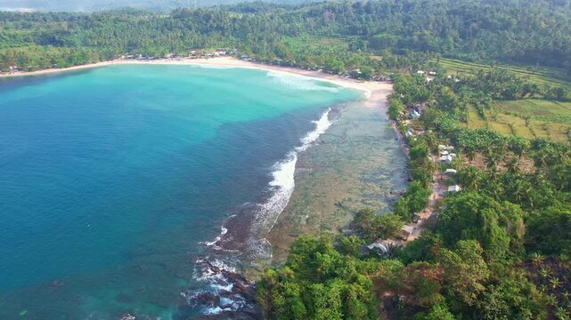 Aerial drone footage of Legon Pari beach, in Lebak Regency, Banten, Java island, Indonesia, with forests around, beautiful clear blue sea and a clean reef, and many food stalls and homestays