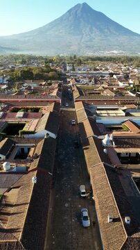 Aerial drone view of a colonial street in Antigua Guatemala aligned with Volcan de Agua rising in the distance. Vertical