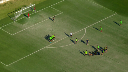 Aerial view of a soccer field during a training session or match. Players in green jerseys are positioned on the grass under bright sunlight, with long shadows stretching across the penalty area. © Stefano Tammaro
