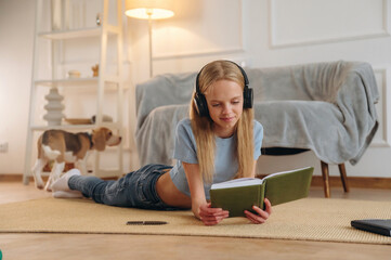 Obraz premium Leisure, in headphones, reading the book. Happy girl is on the floor at home with dog, domestic room