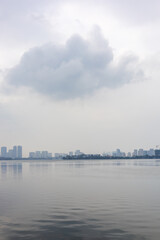 Obraz premium Calm lake with Hanoi skyline in the distance under overcast sky showing urban waterfront, peaceful atmosphere, and minimal cityscape composition with reflections and soft tones