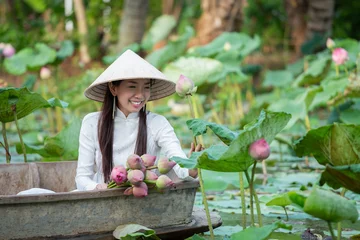 Fotobehang Lotusbloem Southeast Asian woman wearing traditional white dress and conical hat sitting in wooden boat in lotus pond, holding pink lotus flowers, medium shot with soft natural light, peaceful rural lifestyle.  © AU USAnakul