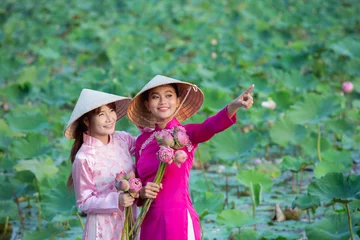 Fotobehang Lotusbloem Southeast Asian women wearing traditional dresses standing in blooming lotus field, holding pink lotus flowers and pointing forward, medium shot with soft natural light, cultural lifestyle.  © AU USAnakul