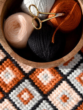 Top view of yarn balls in wooden bowl and vintage scissors on bargello needlepoint blanket, colorful wool thread for embroidery, cozy handmade geometric flame stitch craft background