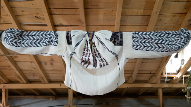 Traditional embroidered Romanian folk blouse hanging on a pole beneath a wooden attic ceiling.