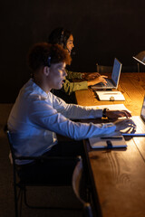 African American colleagues typing on laptops, speaking into headsets at call center desk with lamp