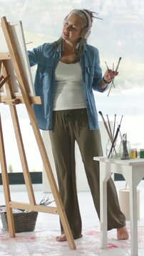 Vertical video: Reaching canvas, artist selecting brushes and painting strokes at easel, headphones