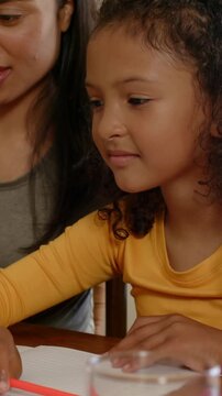 Vertical video: Leaning yellow kid tracing red pencil on paper at table mom guiding hand learning