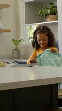 Vertical video: Packing for school girl in yellow placing blue folder into mint backpack in kitchen
