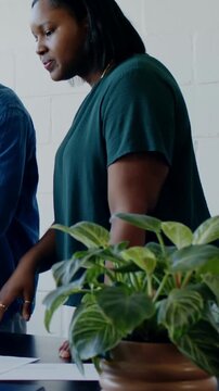 Vertical video: Spotting papers, woman in green top leaning at table, discussing work near plant