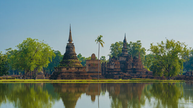 Multiple ancient Buddhist stupas and ruins reflected in a lake with a palm tree at Sukhothai