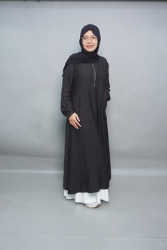 Smiling woman in black hijab and dress stands calmly. She wears glasses and a long-sleeved, floor-length garment. White underskirt adds soft contrast to her dark outfit. Her pose is relaxed, friendly