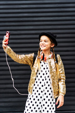Portrait of smiling girl wearing hat and golden sequin jacket taking selfie with smartphone