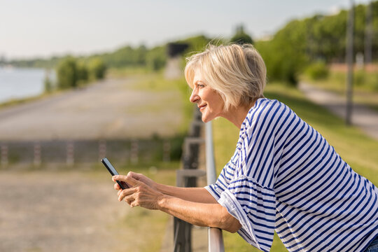 Smiling senior woman with cell phone leaning on railing at riverbank