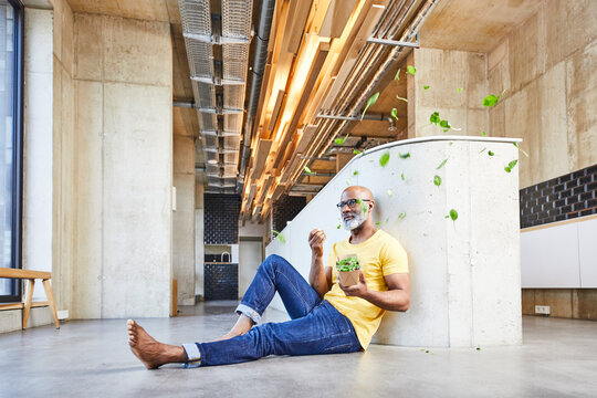 Mature businessman sitting on the floor in modern office surrounded by floating lettuce leaves