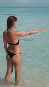 A woman in a bikini stands near the water pointing at a fish swimming below. She seems intrigued by the aquatic creature.
