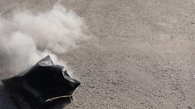 Heat-resistant bag airborne above asphalt as a lithium battery ignites inside, releasing thick smoke. Captures a moment of combustion, motion, and safety testing in an outdoor setting