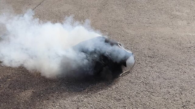 Laptop lithium battery failure inside a heat-resistant safety bag producing visible white smoke, with the protective enclosure containing damage on an asphalt surface