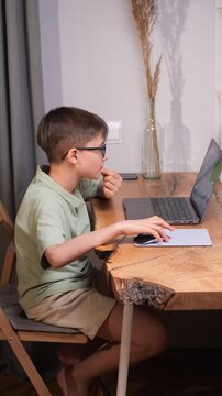 Vertical video young boy wearing glasses working on a modern laptop computer, sitting at a wooden desk with a mouse and mousepad, concentrating on his remote education