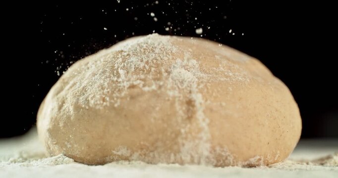 Static shot of a leavened dough loaf isolated on black background with falling flour in super slow motion, 1000 fps. Concept of baking, artisan bread making and dough preparation.