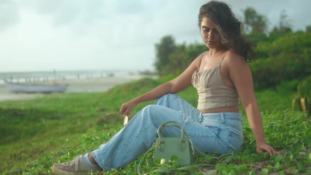 Wamiqa Gabbi sitting on grass near the ocean, relaxing in light denim jeans and a beige crop top, enjoying a casual outdoor fashion moment by the shore