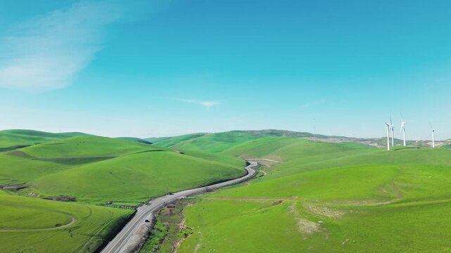 Stunning aerial footage of the vibrant green rolling hills and wind turbines at Altamont Pass Wind Farm in California, showcasing a winding road cutting through the scenic landscape
