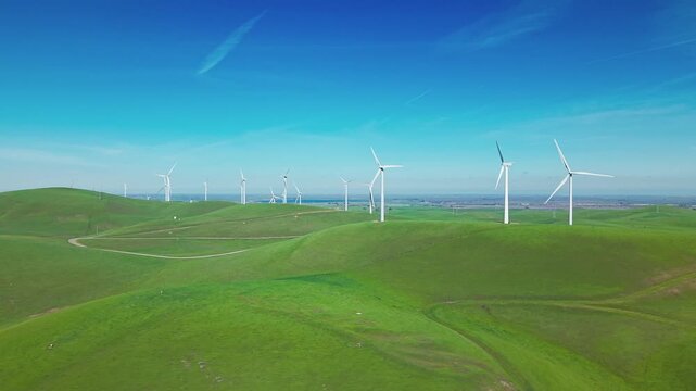 Breathtaking aerial footage over the rolling green hills of the Altamont Pass wind farm in California, with white turbines generating clean renewable energy against a clear blue sky