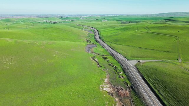 Stunning aerial footage showing a scenic road winding through the lush, green rolling hills of Altamont Pass in California, with vehicles traveling along the empty highway on a sunny day