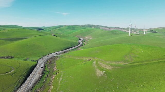 Stunning aerial view of cars driving on a scenic road through the vibrant green hills of Altamont Pass, with modern wind turbines generating clean, renewable energy under a clear blue sky