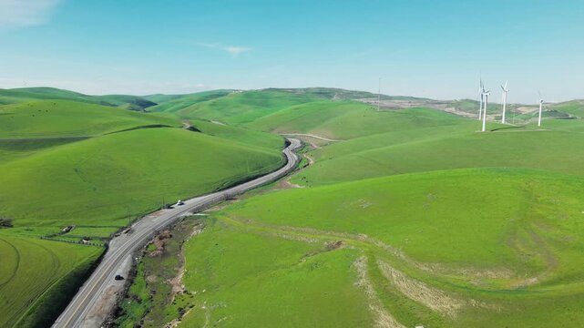 Aerial view of cars driving along a winding road through the vibrant green rolling hills of Altamont Pass, with modern wind turbines generating clean, renewable energy under a clear blue sky