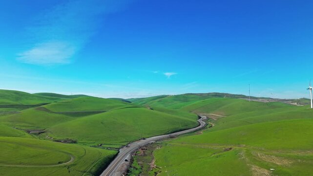 Aerial drone footage flying over the vibrant green rolling hills of Altamont Pass, California, revealing a winding road and distant wind turbines under a clear blue sky on a sunny day