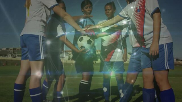 Diverse female players with teenage goalkeeper wearing gloves placing hands on soccer ball on pitch
