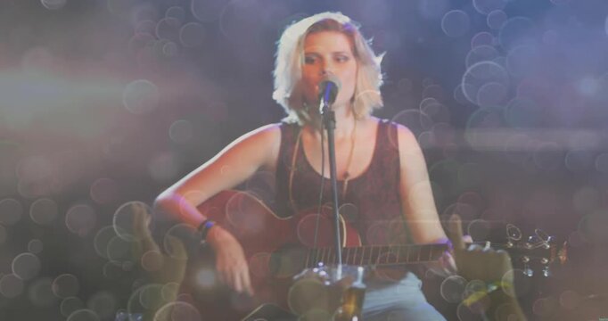 Female singer playing guitar and singing on stage as phones recording music, bokeh framing scene