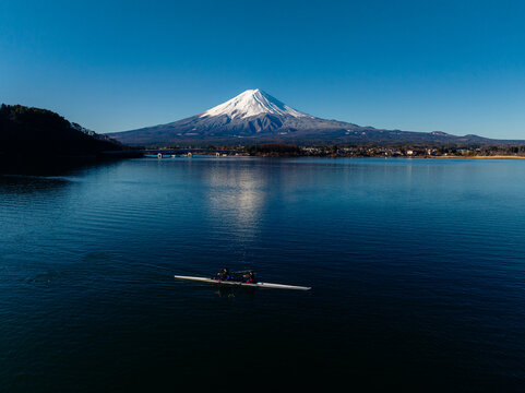 Aerial view of Mount Fuji's snow-capped peak reflected in the serene lake, with a rowing team gliding across the water, Fujiyoshida, Yamanashi, Japan.