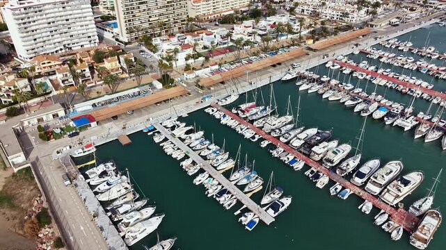 Yachts and boats are docked in the marina while buildings and palm trees line the shore. The scene shows a busy coastal area under a clear sky.