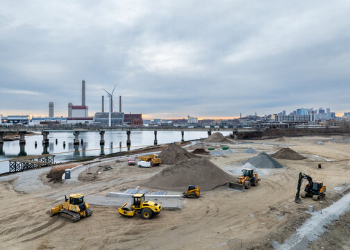 Aerial view of construction vehicles working on the site of a new development near the river, with a power plant and wind turbine in the background. Somerville, Massachusetts, USA.