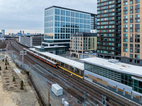 Aerial view of an orange passenger train pulling into Assembly Row station, surrounded by modern architecture. Somerville, Massachusetts, USA.