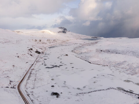 Aerial view of frozen Loch Fada and snow-covered landscape, with the iconic Old Man of Storr piercing the sky under a dramatic cloudy sky, Isle of Skye, Scotland.