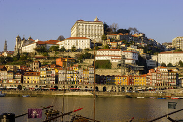 Obraz premium A view of the colorful buildings along the Douro River in Porto, Portugal, with boats docked in the foreground and a large building on the hill behind.