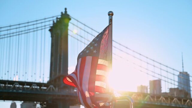 Old shabby American flag flaps in the wind at sunset. Blurred view of the Manhattan Bridge from low angle view.
