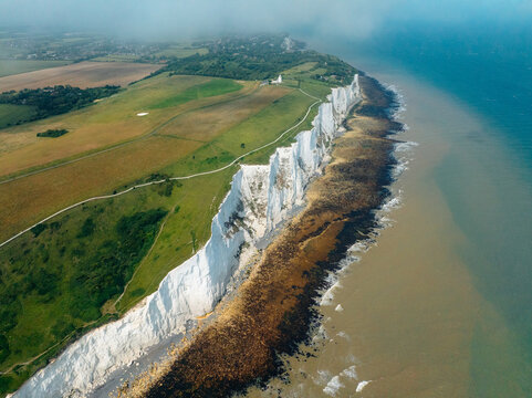 Aerial view of dazzling white cliffs meet the turbulent sea, creating a stark contrast against the verdant fields and the distant, hazy horizon, Dover, England, United Kingdom.
