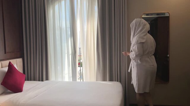 A woman closes the tulle curtains, sits on the bed, a towel on her hair, a robe, and washes her hair at the hotel. High quality 4k footage