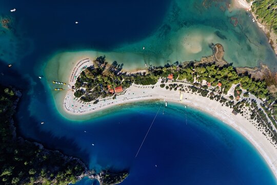 Aerial view of the luminous turquoise waters embracing the serene sandy beach, dotted with colorful umbrellas and lush greenery, Oludeniz, Mugla, Turkey.