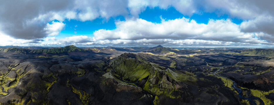 Aerial view of stark, dark volcanic highlands contrasted by vivid green moss and winding glacial rivers under a dynamic sky, Skaftarhreppur, Iceland.