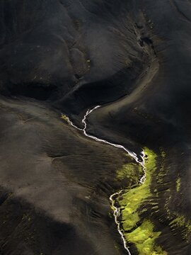 Aerial view of a silver river snaking through a black volcanic landscape, accented by patches of vibrant green moss, Iceland.