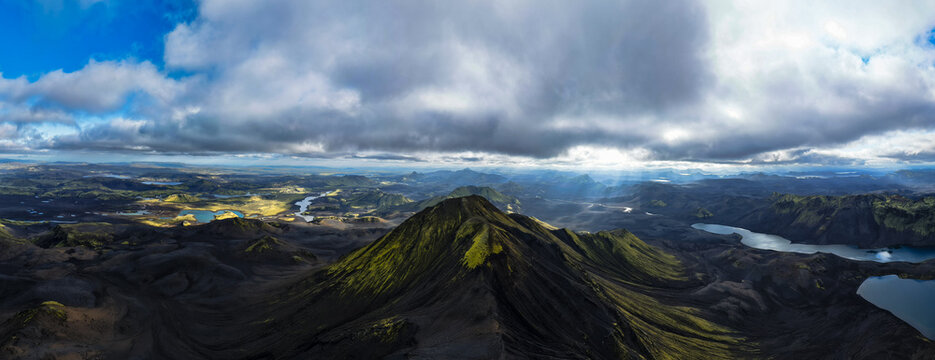Aerial view of sun rays filter through the clouds, illuminating the rugged peaks and still lakes, creating a stunning contrast of light and shadow, Iceland.