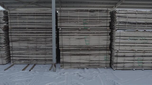 Large stacks of flat panels or boards stored under a metal-roof shelter in a snowy outdoor yard, showing industrial organization and winter conditions