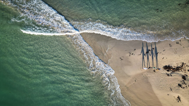 Aerial view of foamy waves crashing on the sandy beach where human shadows stretch long in the afternoon sun, Cleder, France.