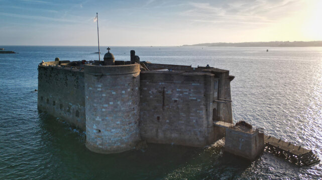 Aerial view of Chateau du Taureau stone fortress standing majestically in the sea, the water reflecting the soft sunlight, creating a serene yet formidable vista, Plouezoc'h, Bretagne, France.